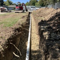 PVC sewer pipe installation with trenching for plumbing repair or new construction. Heavy equipment and fresh dug earth indicate ongoing underground pipeline work.
